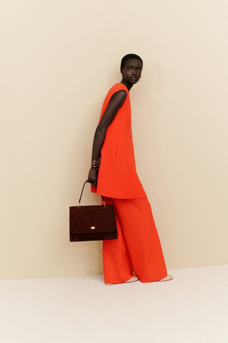 A woman stands in a studio wearing a red sleeveless top and wide-legged pants, carrying a brown square handbag, against a neutral backdrop.