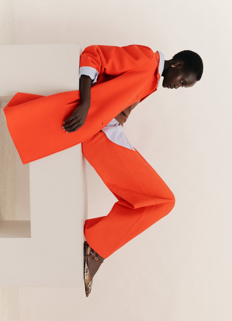 A tall, slender fashion model poses wearing an orange pants suit with patterned flats against a cream colored wall in studio lighting.