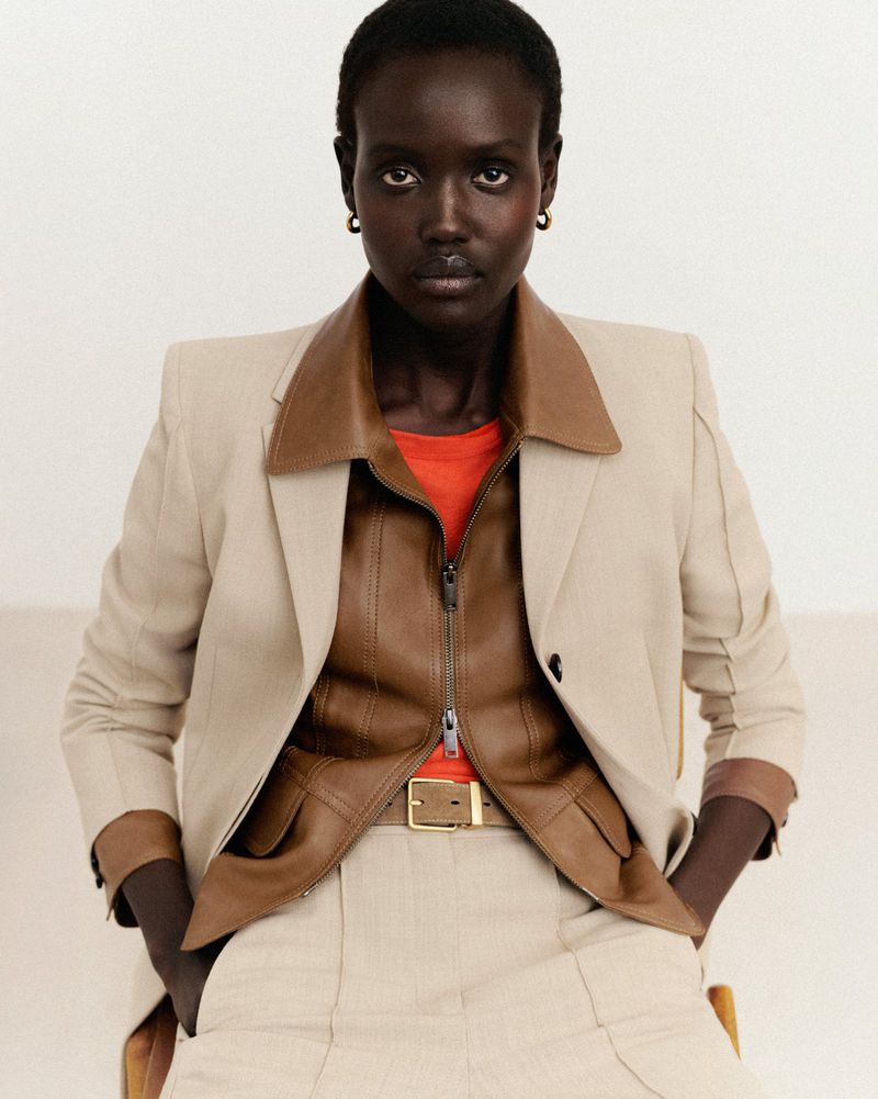 Portrait of an adult woman in a blazer, orange top, and brown leather vest with a beige belt. She has dark skin and dark hair.