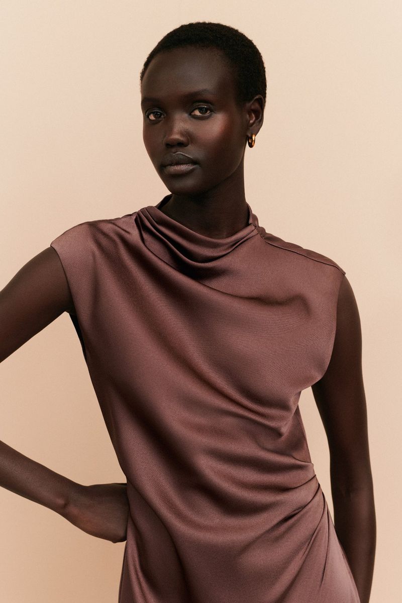 Portrait of a dark-skinned Black woman in a brown sleeveless satin dress, posing indoors against a neutral background with subtle studio lighting.