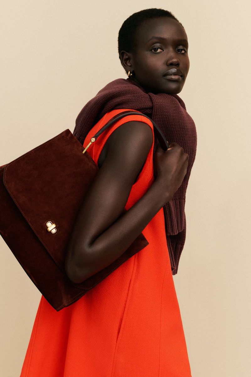 Dark-skinned woman in an orange dress and maroon sweater poses with a brown handbag against a neutral backdrop in a fashion studio shoot.