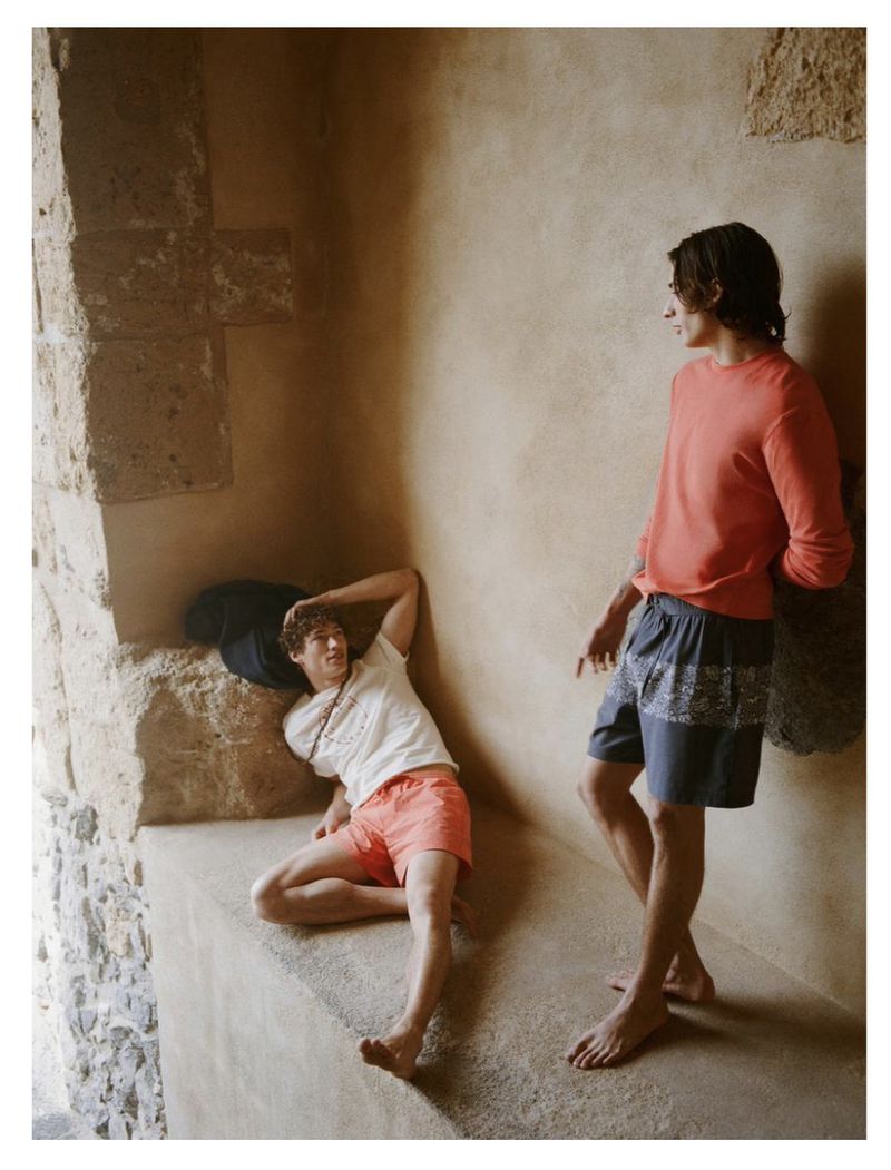 Two young adult males in casual summer clothing pose indoors against a textured wall; one reclines, and the other stands barefoot.