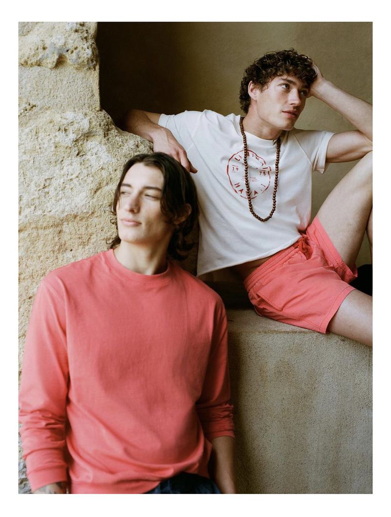 Two adult male fashion models pose on a weathered stone structure in a medium shot. The man on the left wears a pink long-sleeved shirt.