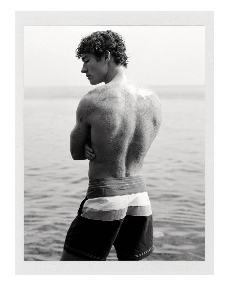 A man with curly hair wearing colour-block swim shorts stands with his back to the camera in front of a calm, vast ocean.