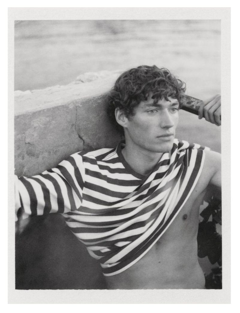 Monochrome portrait of a young adult male with curly hair, partially undressed in a striped top, leaning against a weathered stone structure.