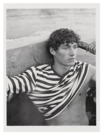 Monochrome portrait of a young adult male with curly hair, partially undressed in a striped top, leaning against a weathered stone structure.