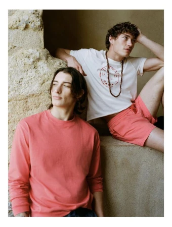 Two male models posing against a textured stone wall wearing coral and white summer apparel for Zara