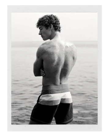 Monochrome portrait of a shirtless male model wearing striped swim trunks standing by the water for Zara Man.
