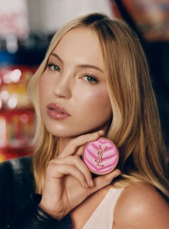 Close-up of a blonde woman holding a YSL makeup compact, wearing a black jacket and a white top, indoor setting.