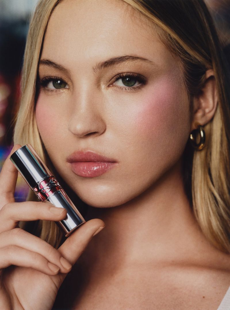 Close-up of fair-skinned woman holding a silver and pink lip product with a blurred background, fair skin, and gold earrings.