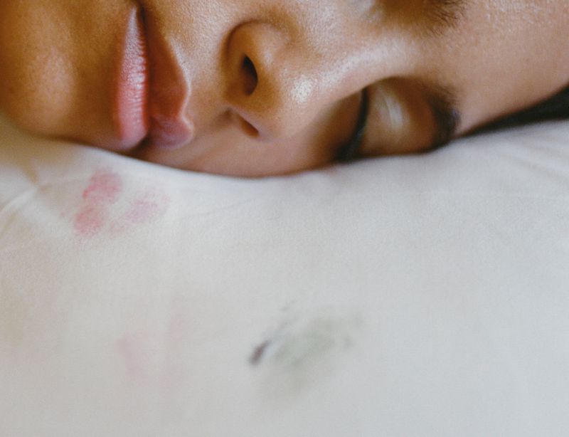 Close-up of a sleeping adult with dark hair, resting on a white pillow with lipstick and face-oil stains, capturing an intimate moment.