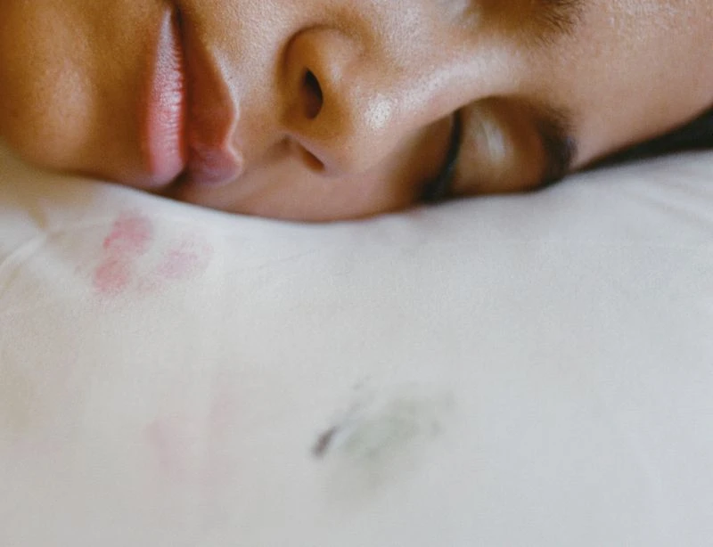 Close-up of a sleeping adult with dark hair, resting on a white pillow with lipstick and face-oil stains, capturing an intimate moment.