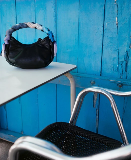 Leather handbag with a braided fabric handle sits on a white table with metal legs, with a blue wooden wall in the background.