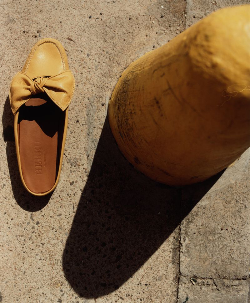 Tan leather mule shoe with bow detail on concrete surface next to yellow pillar casting a shadow. Product shot for fashion blog.