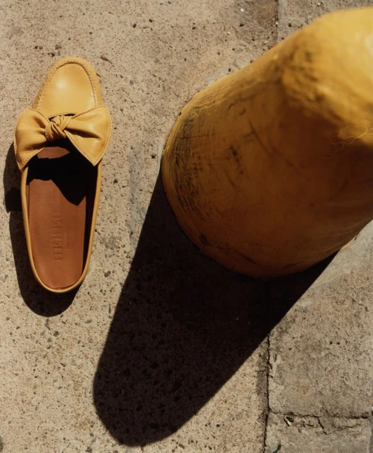 Tan leather mule shoe with bow detail on concrete surface next to yellow pillar casting a shadow. Product shot for fashion blog.