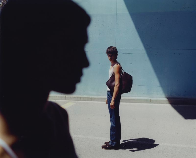 A man in a white tank top and blue jeans standing outdoors next to a dark blue wall carrying a large dark leather tote bag