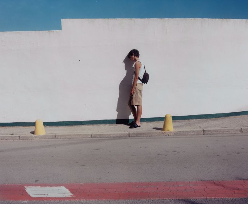 A person stands beside a white wall and yellow traffic cones on a sunny day, casting shadows on the wall in an outdoor setting.