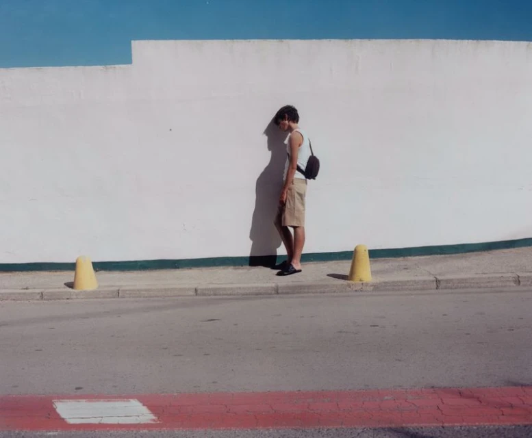 A person stands beside a white wall and yellow traffic cones on a sunny day, casting shadows on the wall in an outdoor setting.