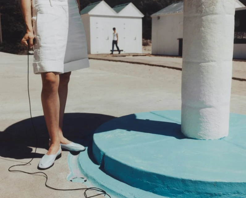 Woman in white skirt and shoes standing near a pool with a person skateboarding near white buildings in the background on a sunny day.