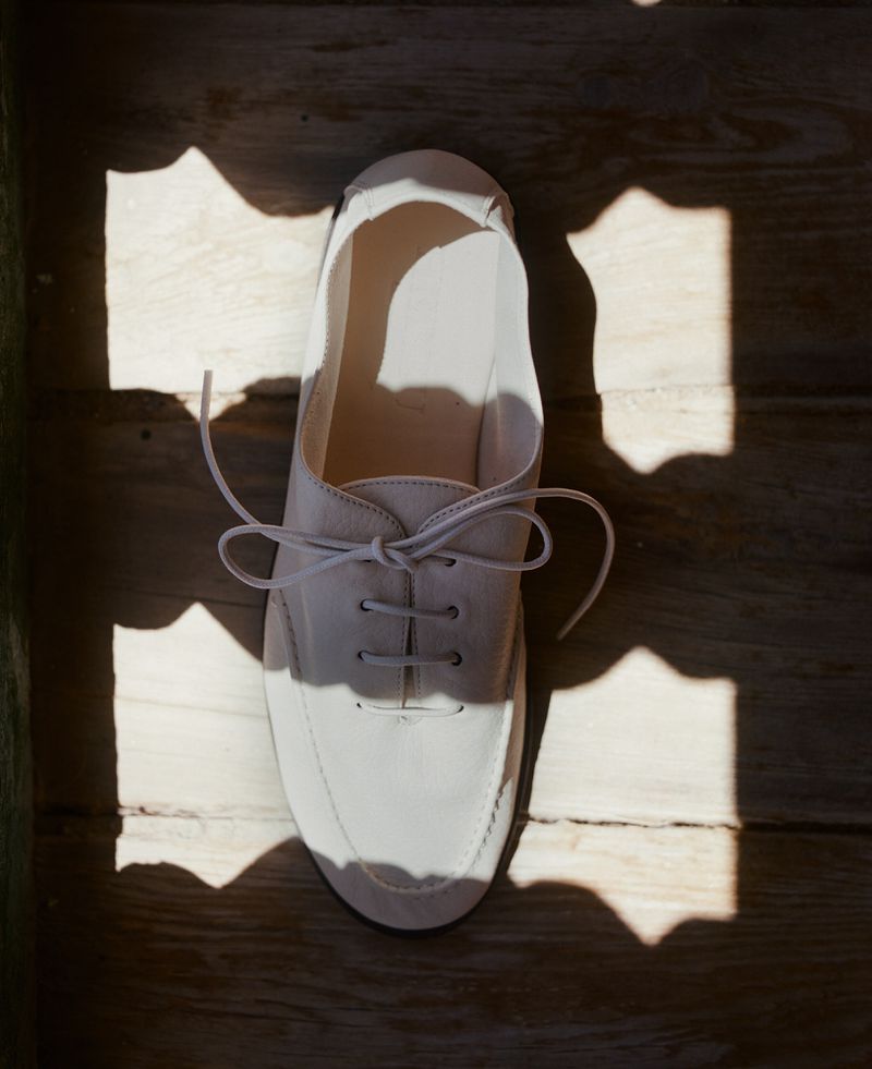 Overhead shot of a white lace-up shoe sitting on wood floor, partially lit by sunlight shining through a patterned window.