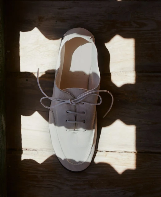 Overhead shot of a white lace-up shoe sitting on wood floor, partially lit by sunlight shining through a patterned window.
