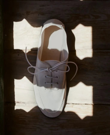Overhead shot of a white lace-up shoe sitting on wood floor, partially lit by sunlight shining through a patterned window.