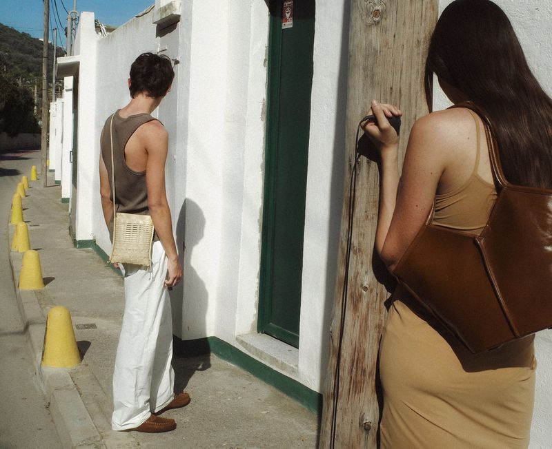 Two people on a street with white buildings. A man in white pants carries a bag. A woman wears a brown dress and carries a brown bag.