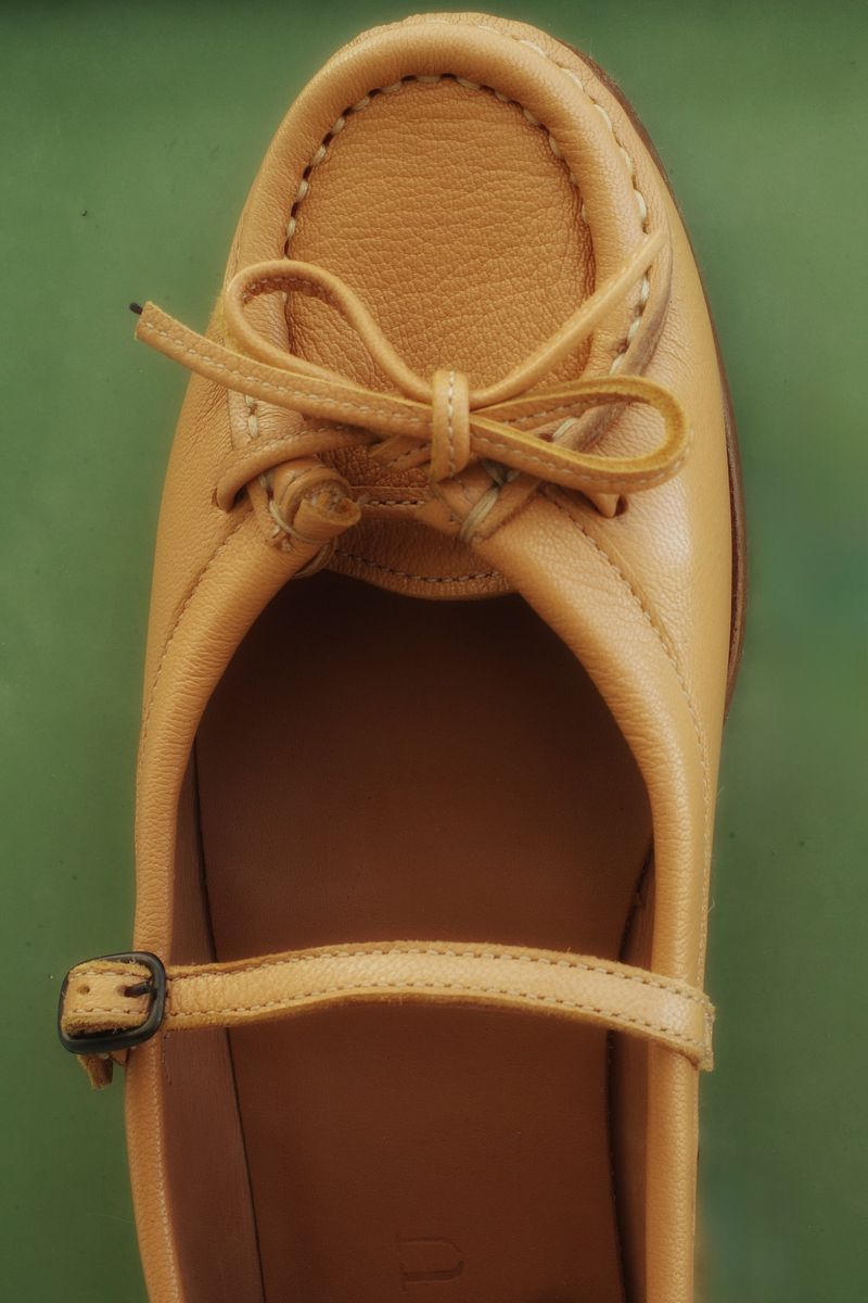 Close-up studio shot of a tan leather Mary Jane shoe with a bow, featuring a buckle strap and moccasin-style stitching, set against a green background.