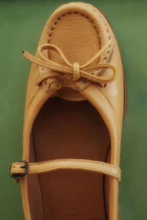 Close-up studio shot of a tan leather Mary Jane shoe with a bow, featuring a buckle strap and moccasin-style stitching, set against a green background.