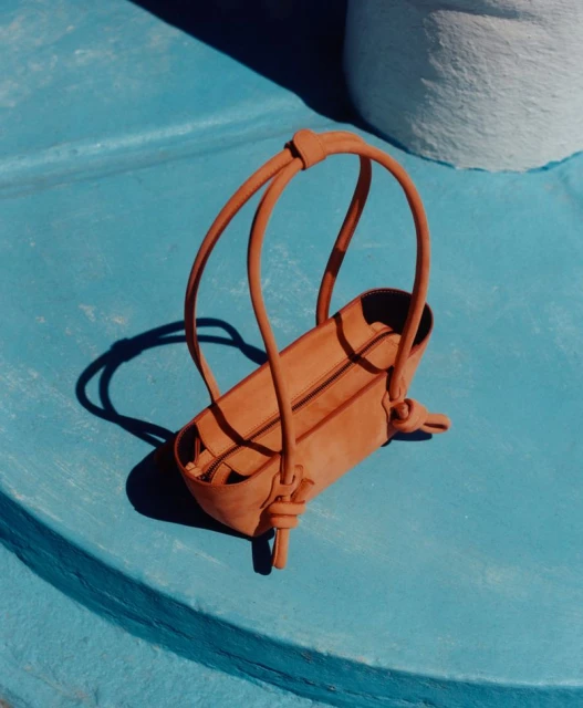 Tan leather purse with unique loop handles on a blue surface, illuminated by natural light, casting sharp shadows and highlighting its texture.