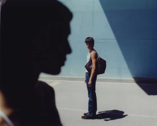 A man in a white tank top and blue jeans standing outdoors next to a dark blue wall carrying a large dark leather tote bag