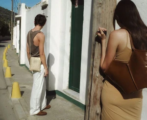 Man in grey tank top and white trousers with woman in tan dress holding a brown leather bag outdoors on a narrow street.