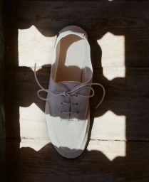 Top-down view of a single off-white leather lace-up shoe resting on wooden slats with harsh overhead geometric shadows.