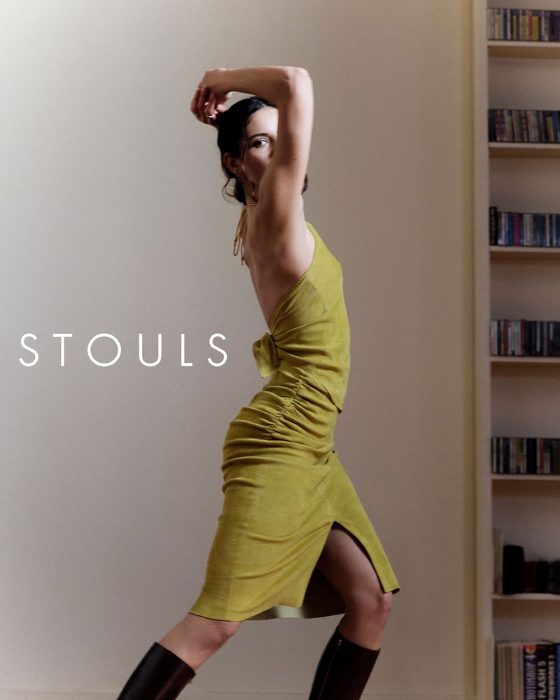 Model in olive green dress and tall boots posing in an interior space with a bookshelf in the background for Stouls Fall Winter 2026