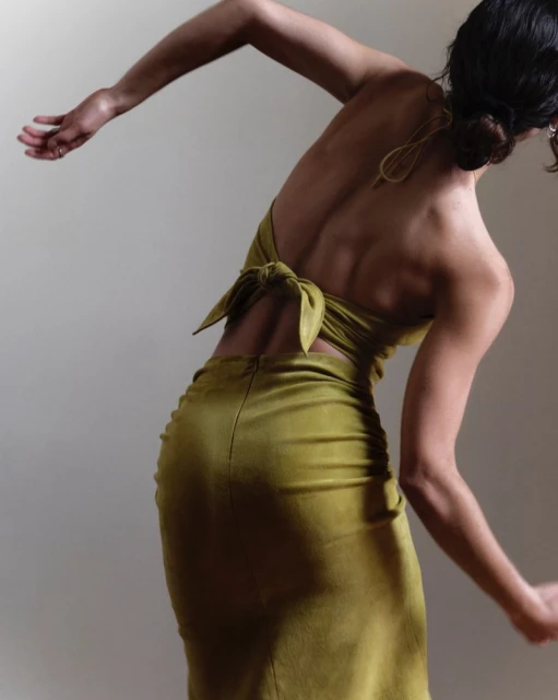 Woman in a chartreuse open-back dress posing in a studio. The model is captured from behind in a three-quarters shot against a plain wall.