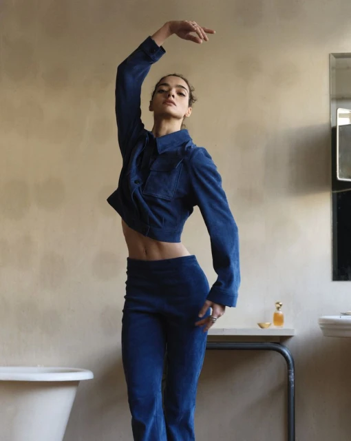 A slender woman in a blue corduroy outfit strikes a pose in a minimalist bathroom setting, with a focus on fashion and elegance.