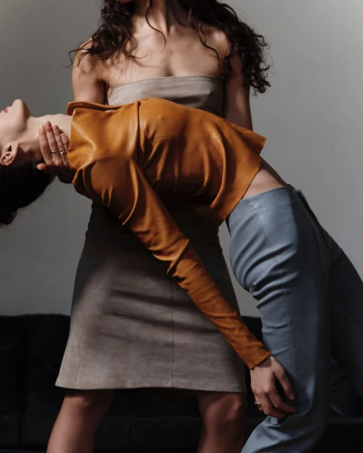 Two women in a studio setting, one in a taupe tube top and skirt, the other in a mustard-colored top and blue pants, fashion shot.