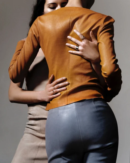 Two women embrace indoors, one in a tan dress, the other in a brown leather jacket and grey pants, showing fashion and connection.
