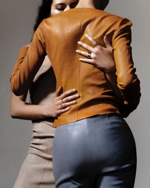 Two women embracing in a fashion campaign with one wearing a tan leather jacket and grey trousers against a neutral background.