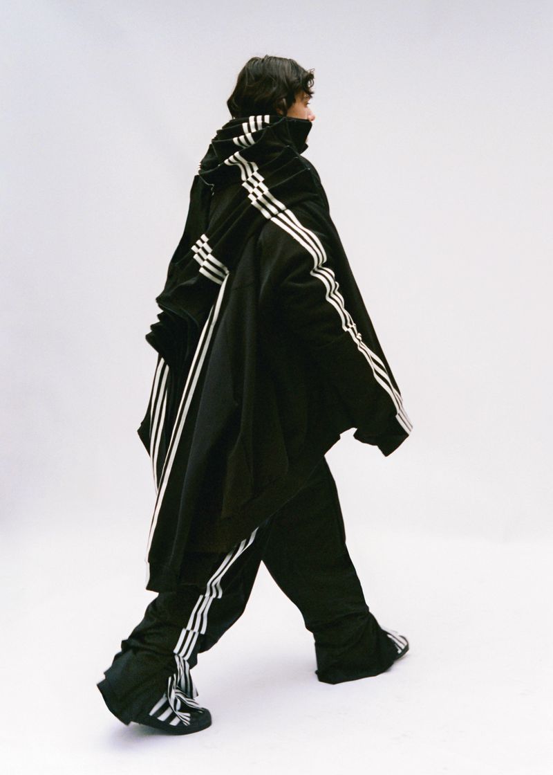 Person in oversized black Adidas tracksuit with white stripes walks against a plain white backdrop in a studio setting.