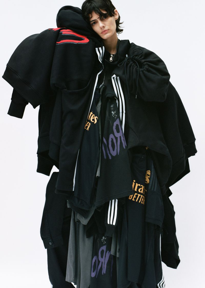 Adult female with short dark hair posing indoors, draped with multiple black tops and shirts, against a bright white backdrop. Fashion shoot.