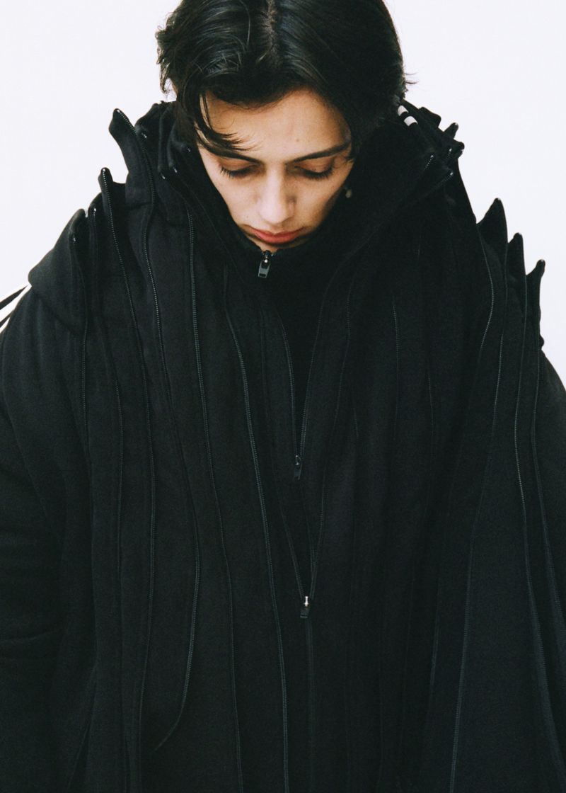 Person in a black, hooded, sculptural coat looking down in a studio setting with a white background. Fashion shot, medium framing.