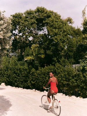 Model Melodie Monrose in a red one-piece swimsuit riding a bicycle along a sandy path lined with lush green trees.