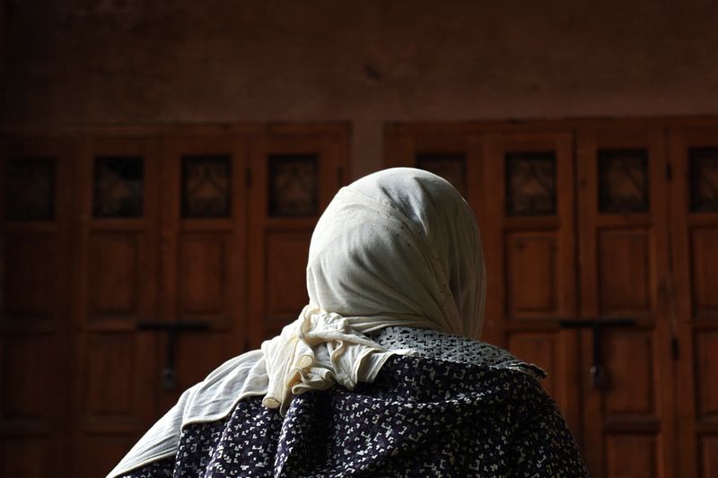 Back view of adult female in headscarf and patterned top, standing indoors with warm lighting in front of a weathered wooden door.