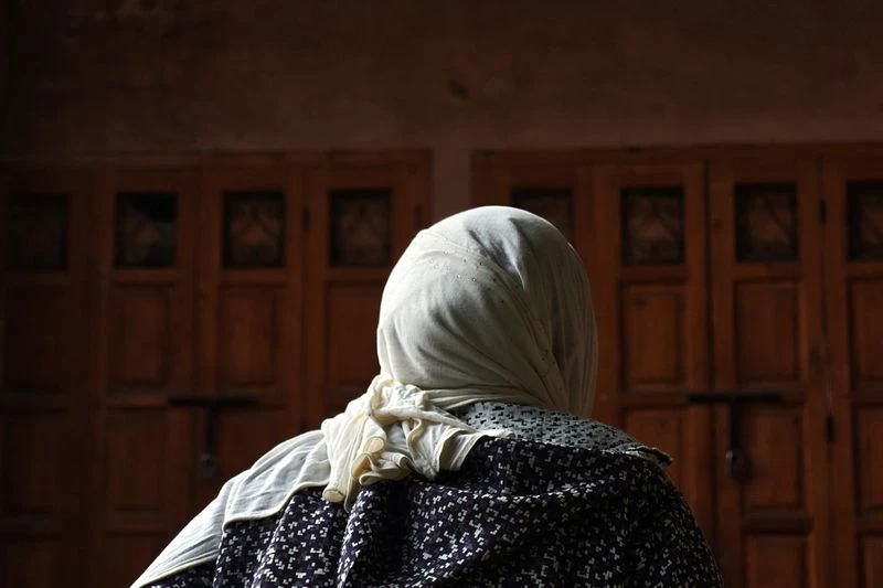Back view of adult female in headscarf and patterned top, standing indoors with warm lighting in front of a weathered wooden door.