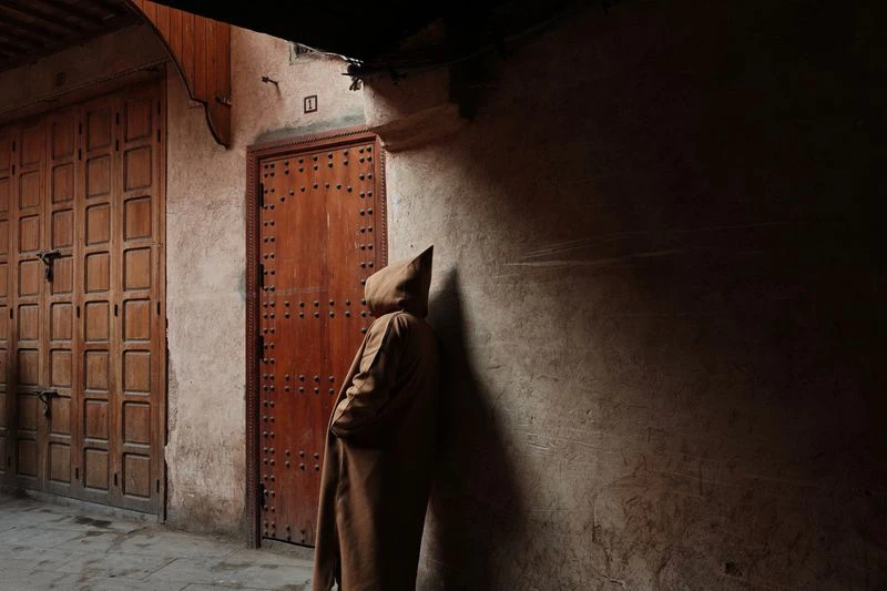Person in hooded robe standing in a dark alleyway with wooden doors and textured wall in dim lighting creating shadows.