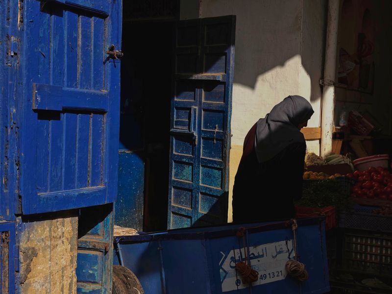 Woman in hijab stands at outdoor market with vibrant blue doors and a cart filled with goods, bathed in warm sunlight.