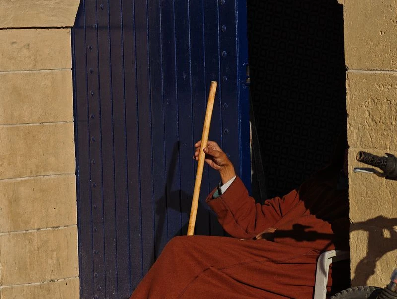 An elderly person sits in a doorway next to a blue door, holding a walking stick, wearing an orange robe, in an outdoor setting.