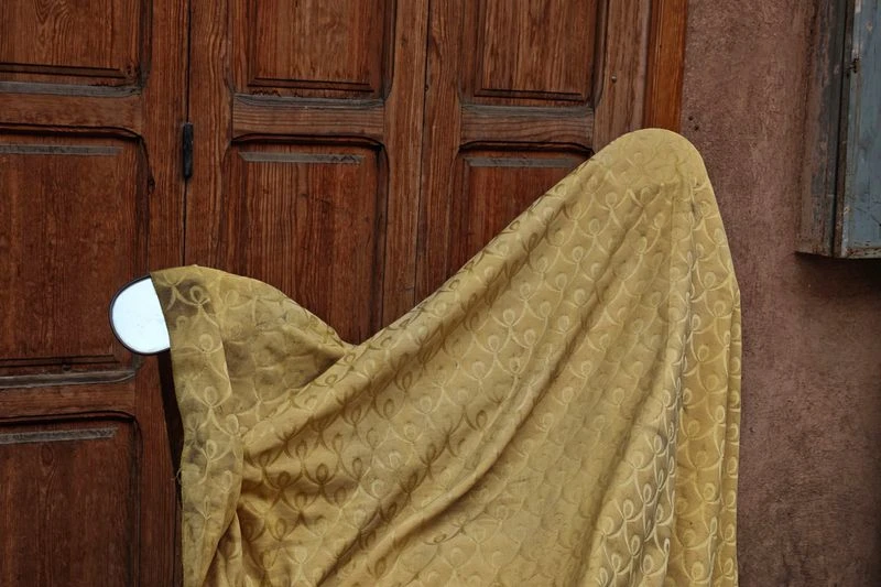 Yellow patterned fabric drapes over a chair, set against a brown wooden door and partially visible stucco wall with a blue window frame.