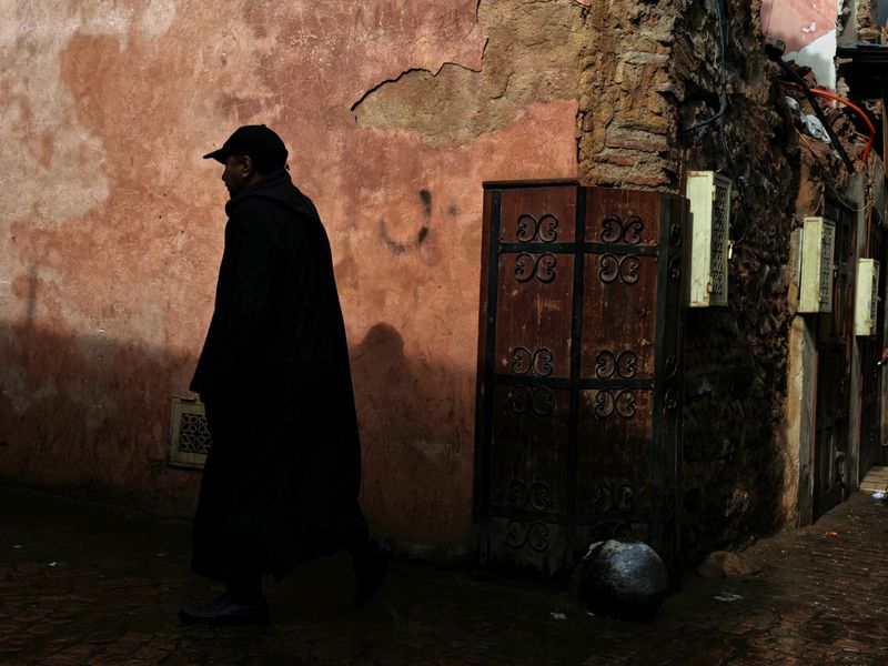Man in dark robe walks along a narrow city street in shadow, with textured walls and an arched doorway and gate visible in background.